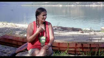 Shelly Boyd - une femme Sinixt avec deux longues tresses noires, vêtue d'un débardeur orange. Elle est assise sur une bûche devant une rivière, avec un canot derrière elle.