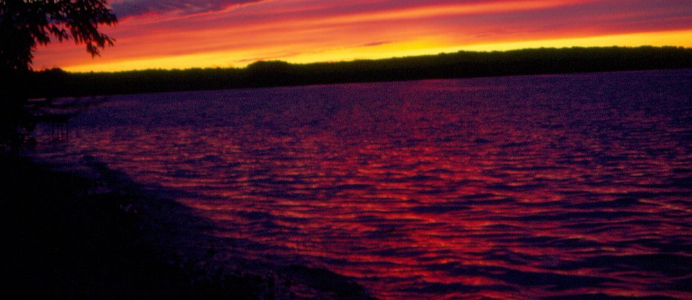 Photographie en couleurs d'un coucher de soleil sur un lac, le ciel prenant des teintes de jaune, d’orange et de mauve.