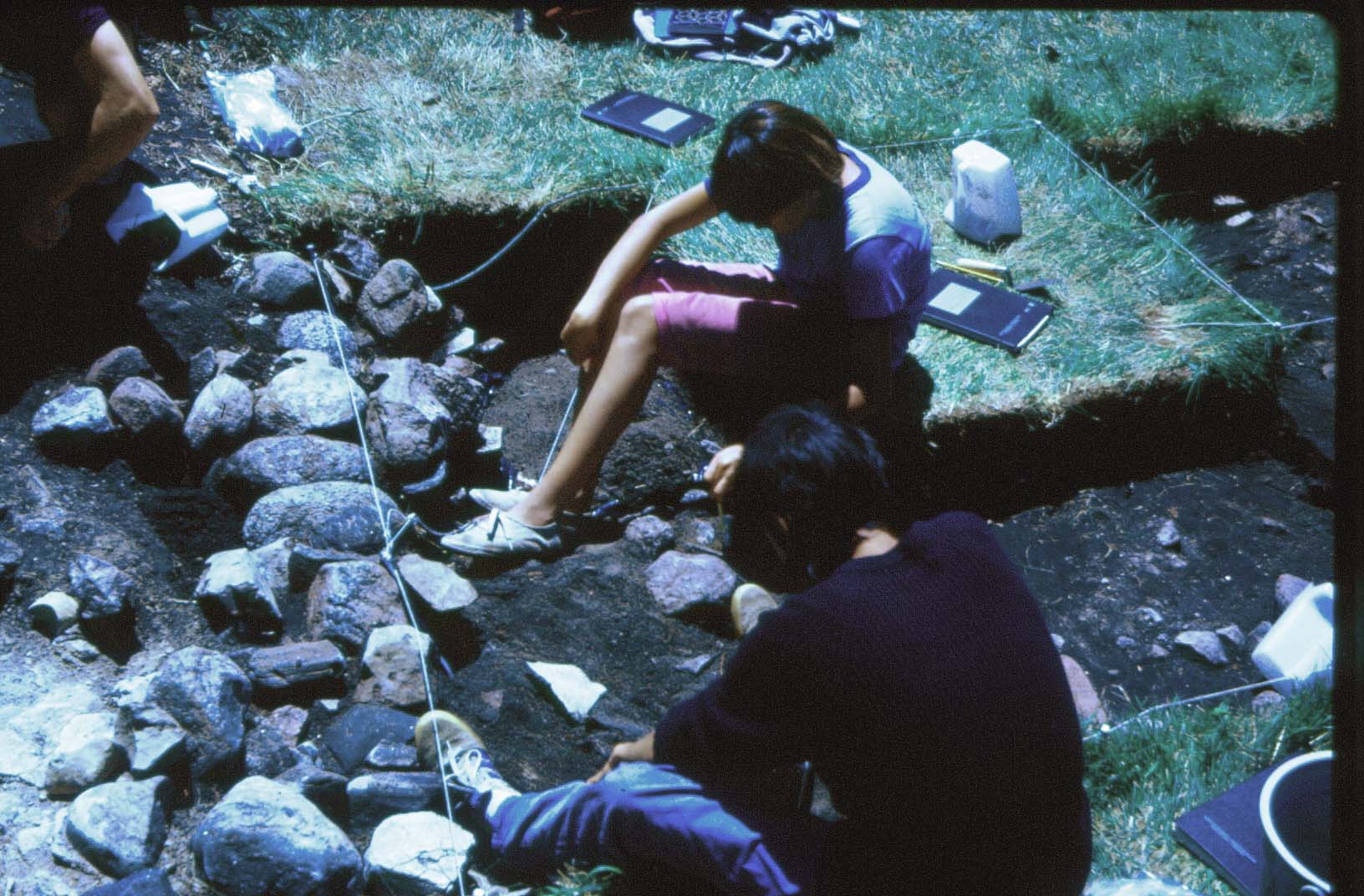  Photographie de deux étudiants en train de fouiller près des murs du Fort. La couche arable a été enlevée et ils sont à peu près à 10 cm en dessous du niveau du sol.