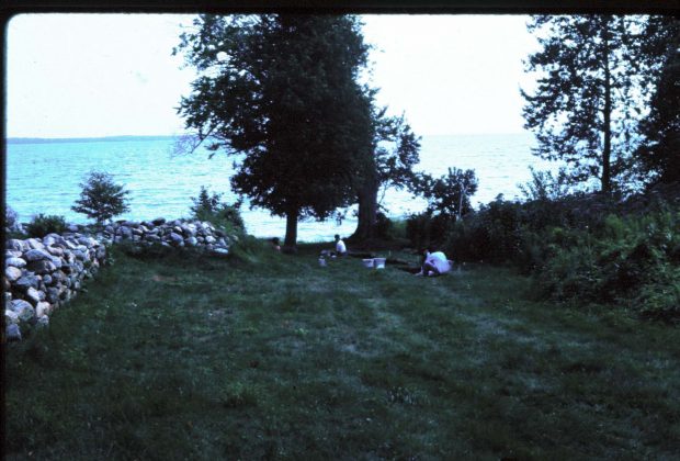 Photographie d'un mur en pierres bas, en forme de L, dans un champs avec le lac en arrière-plan