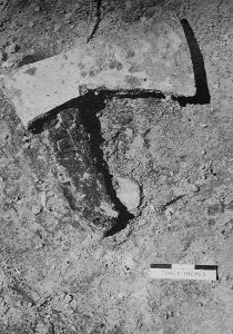 Photographie dans les tons gris d’une hache en fer avec manche calciné, avant son prélèvement.