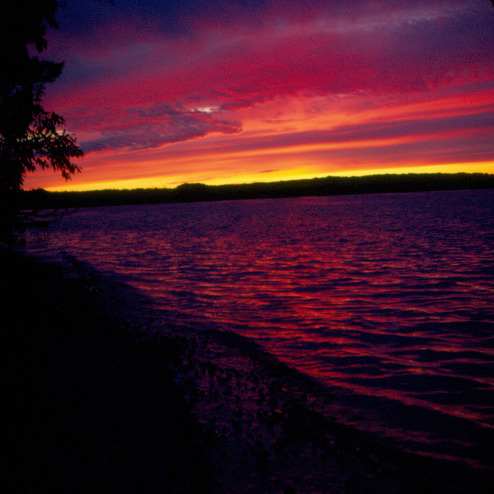 Photographie en couleurs d'un coucher de soleil sur un lac, le ciel prenant des teintes de jaune, d’orange et de mauve.