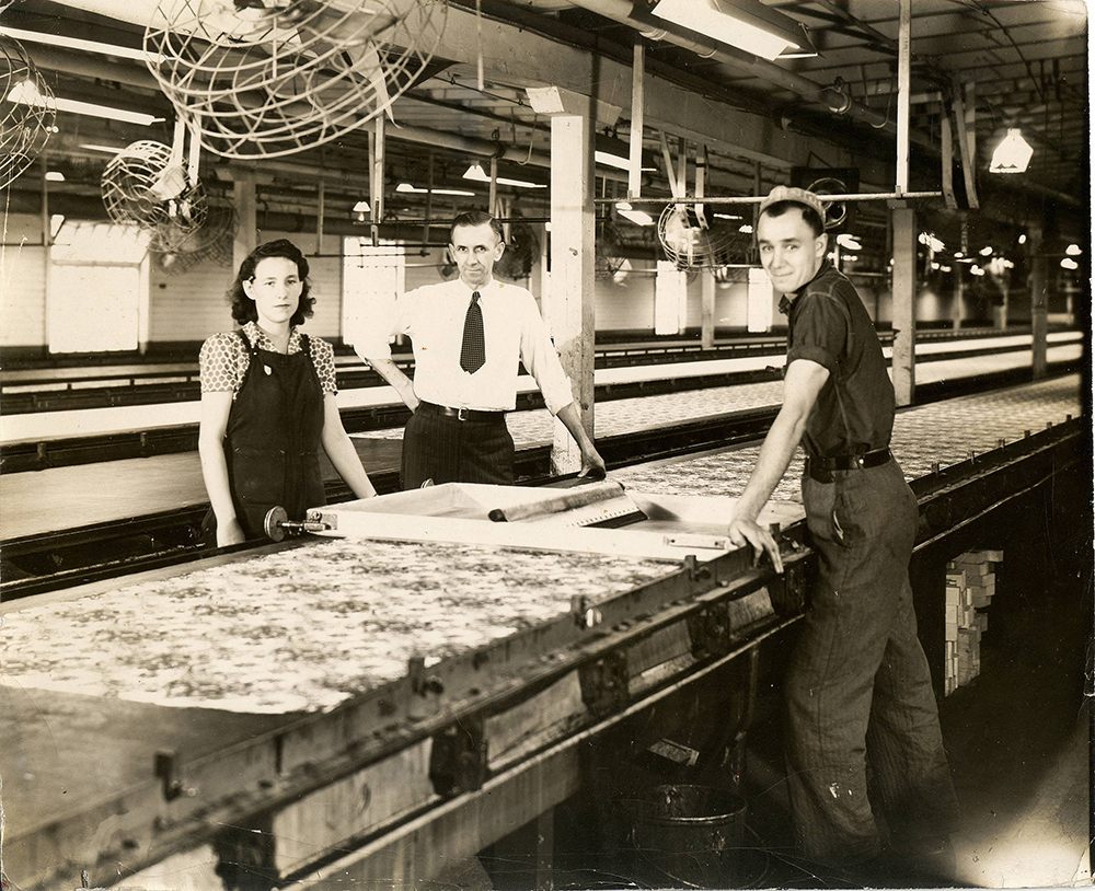 Trois employés, un homme en chemise blanche et cravatte, un homme en habit de travail une femme portant un tablier, posent devant une longue table dans une usine.