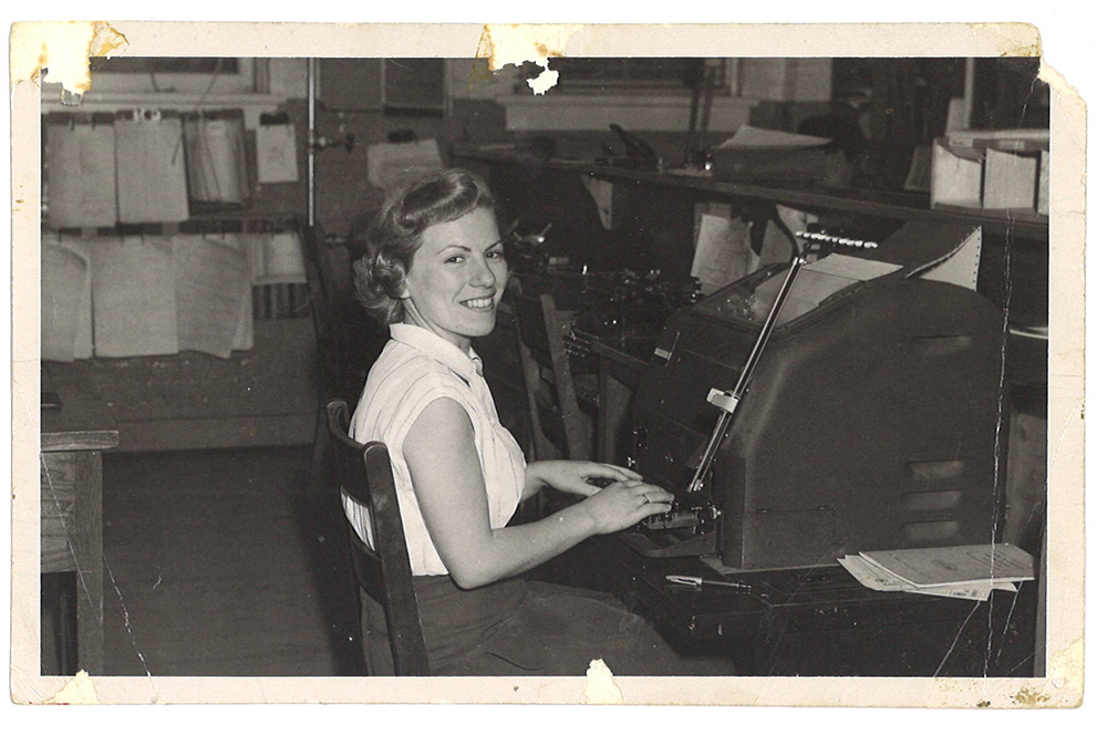 Photo noir et blanc d’une jeune fille assise devant un appareil avec les mains sur un clavier ressemblant à une machine à écrire.