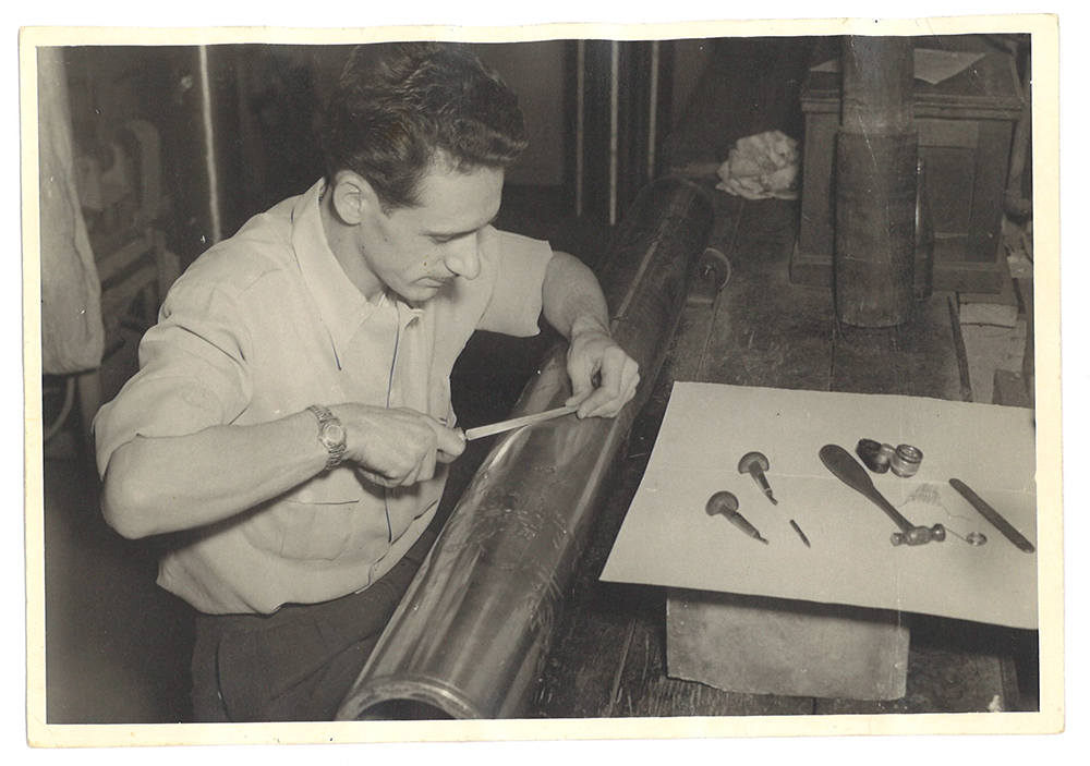 Un ouvrier se penche sur un rouleau de métal avec des petits outils de graveur autour de lui.