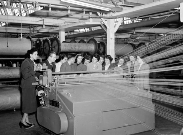 Un groupe de jeunes filles rassemblées devant devant un appareil de tissage à l’intérieur de l’usine