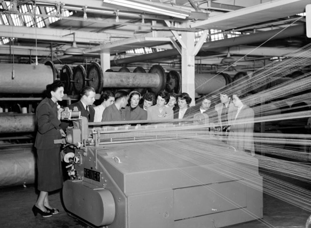 Un groupe de jeunes filles rassemblées devant devant un appareil de tissage à l’intérieur de l’usine