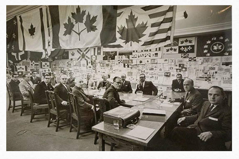 Photo noir et blanc avec 15 hommes sont assis à une longue table avec plusieurs drapeaux suspendus au-dessus d’eux.