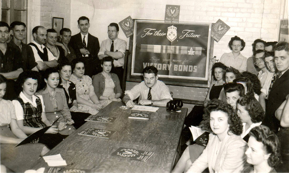 Un groupe de 28 employés autour d’une table, femmes et hommes, posent lors d’une séance de promotion pour l’achat de bons de la victoire en 1945.