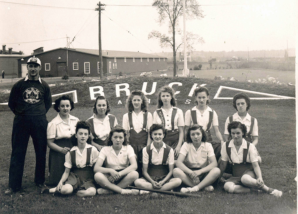 Équipe de balle féminine et de leur entraîneur sur deux rangs devant un talus sur lequel est inscrit « Bruck » avec des bâtiments à l’arrière-plan.