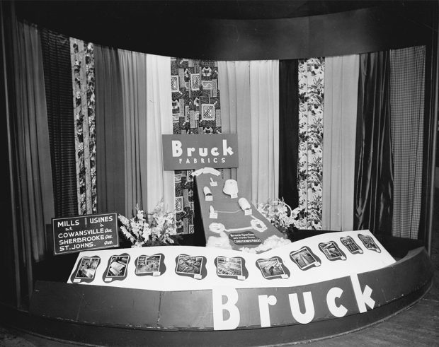 Kiosque de promotion des tissus Bruck avec une enseigne identifiant les usines de Cowansville, Sherbrooke et St-Johns. Des draperies, des outils de fabrications et des photos des installations de la compagnie sont aussi en démonstration. 