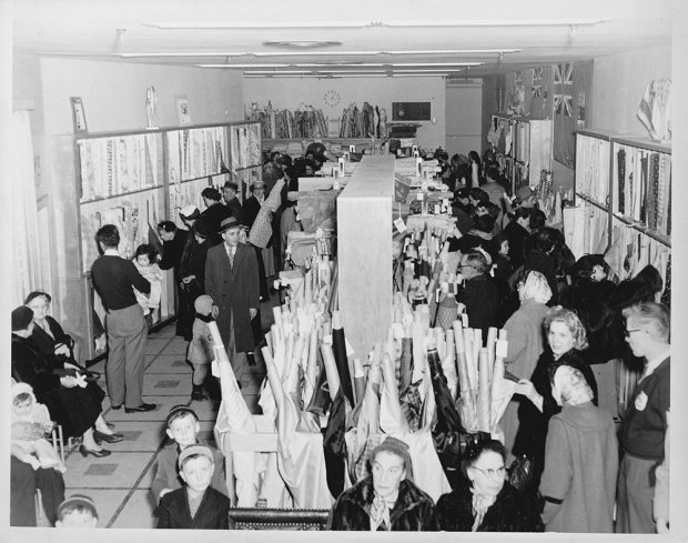 Photo noir et blanc montrant plusieurs clientes et clients autour de rouleaux de tissus dans le magasin de tissus de la Bruck.