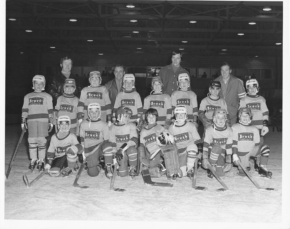 Équipe de hockey mineur de la Bruck Silk Mills avec les entraîneurs sur la patinoire d’un aréna, photographiés sur trois rangs.