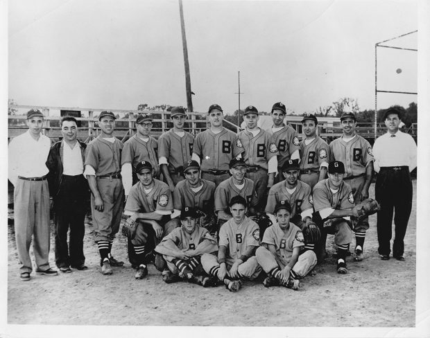 L’équipe de baseball Bruck en uniforme sur trois rangs avec les entraîneurs, devant un terrain de baseball.