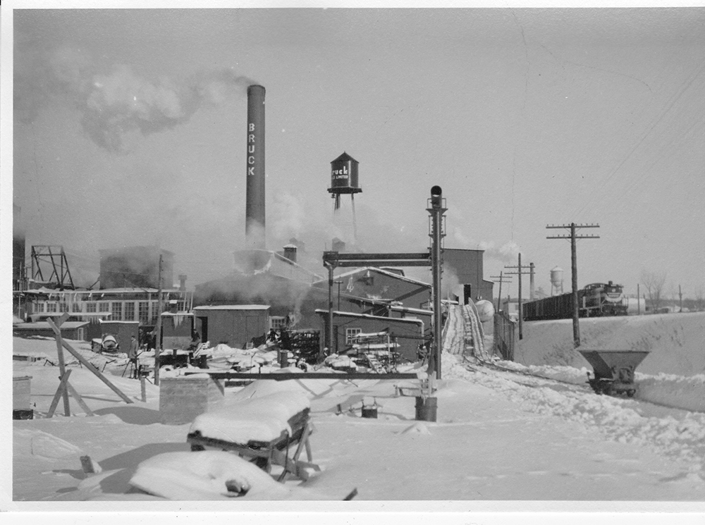 Photo noir et blanc de l’usine Bruck  l’hiver avec un train qui passe à proximité à droite.