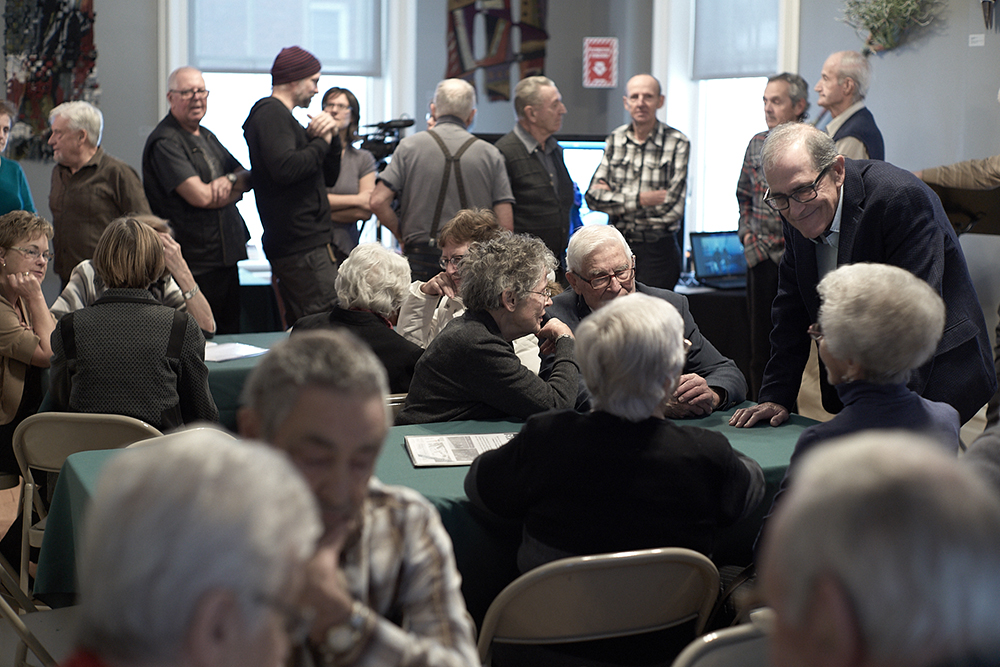 Photo en couleur où des personnes âgées sont assises autour de plusieurs tables à l’avant-plan et discutent. D'autres personnes sont debout à l’arrière-plan et conversent en petits groupes devant deux fenêtres et un écran.