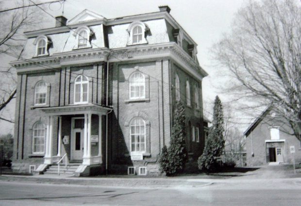 Photo noir et blanc d’un bâtiment en brique avec des colonnes en avant et huit fenêtres à lucarne. Il y a un plus petit bâtiment à droite à l’arrière avec un toit pointu.  