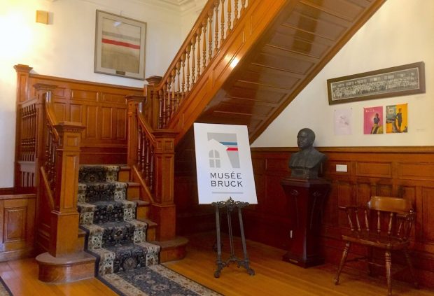 Photo d’un hall d'entrée avec un escalier en bois, une toile encadrée, une affiche sur un chevalet et un buste en bronze.