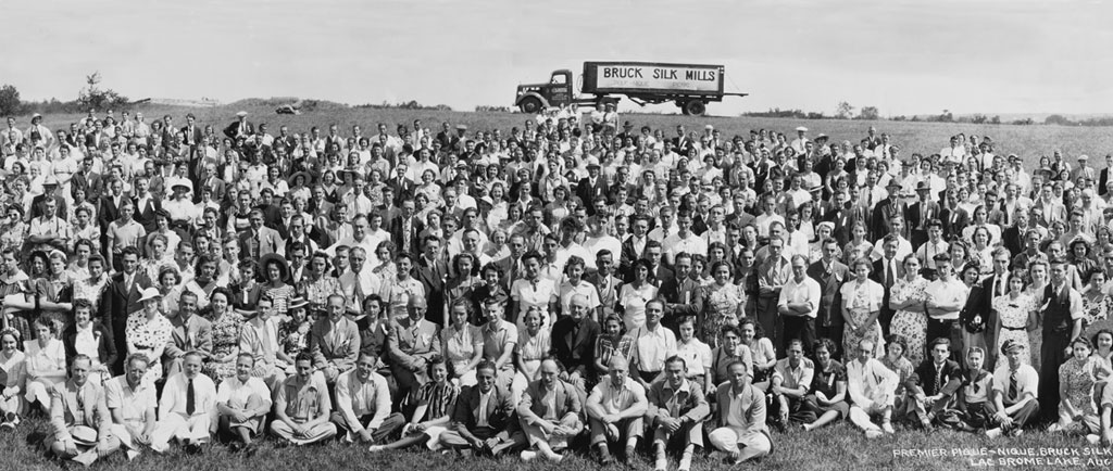 Photo de groupe en noir et blanc avec plusieurs rangées d’hommes et de femmes qui sont assis ou debout dans un champ et qui regardent la caméra. Un camion stationné apparaît au loin, en haut de la photo.