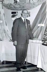 Gerald Bruck, wearing glasses and a suit poses in front of a table where fabrics are draped all around