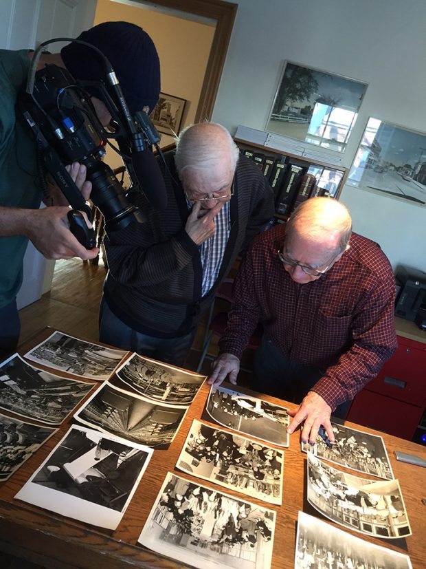 Un caméraman filme deux hommes âgés qui regardent des photographies placées sur une table.