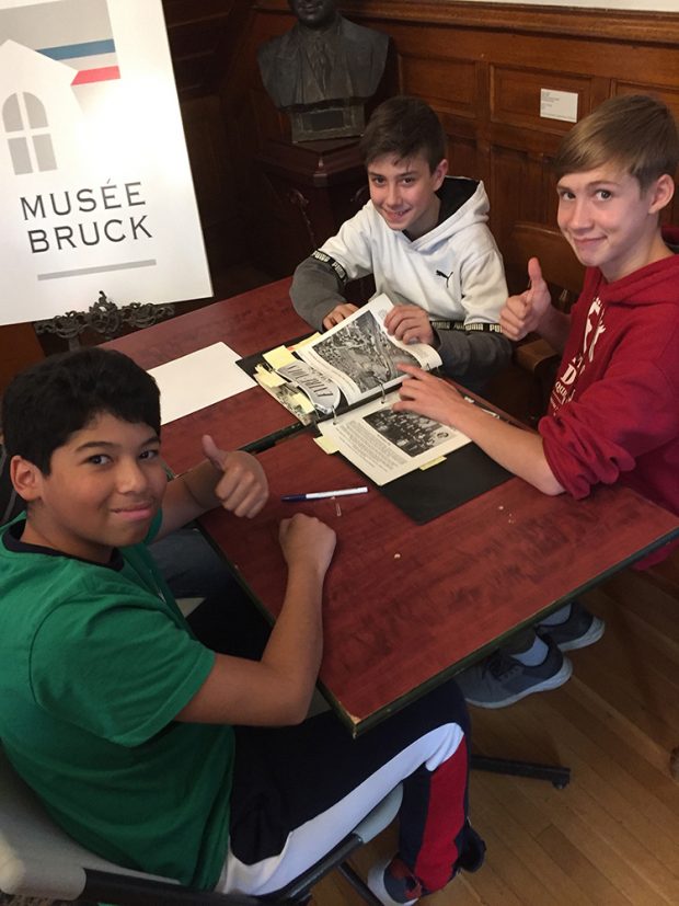 Trois jeunes garçons attablés devant un cartable avec une affiche du logo du Musée Bruck et un buste en bronze à l’arrière-plan.