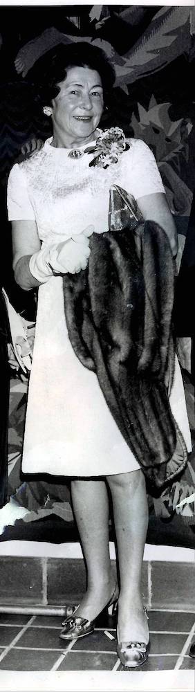 Portrait noir et blanc d’une dame souriante (Eugenie Sharp Lee) tenant un petit sac à main et un manteau de fourrure sous son  bras.