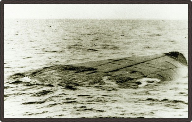 Photo en noir et blanc d’un navire chaviré flottant sur l’eau. On n’aperçoit qu’une portion de la coque du navire.