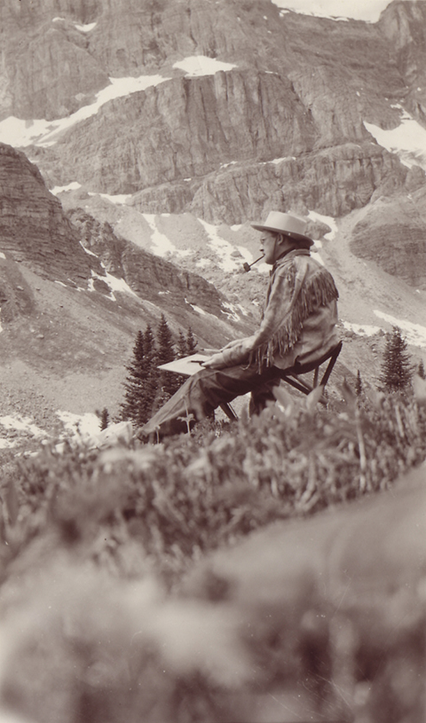 Photo sépia d’un homme portant une veste en daim à franges, un chapeau et fumant la pipe, peignant dans un paysage de montagne.