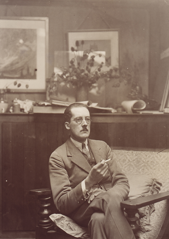 Portrait sépia d’un homme moustachu assis dans une pièce à boiseries, portant lunettes, costume et cravate et fumant une cigarette. Tableaux sur les murs.