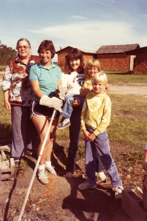 Photo couleur de trois adultes et deux enfants, avec des dépendances en arrière-plan.