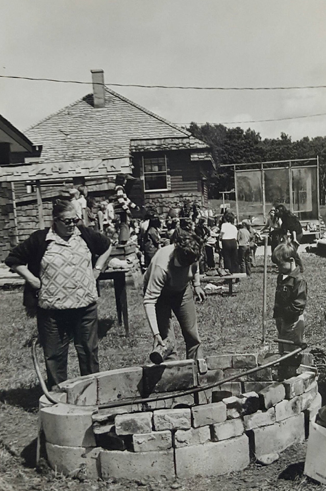 Photo noir et blanc de deux adultes et d’un enfant autour d’un feu de camp avec l’ancienne école derrière.