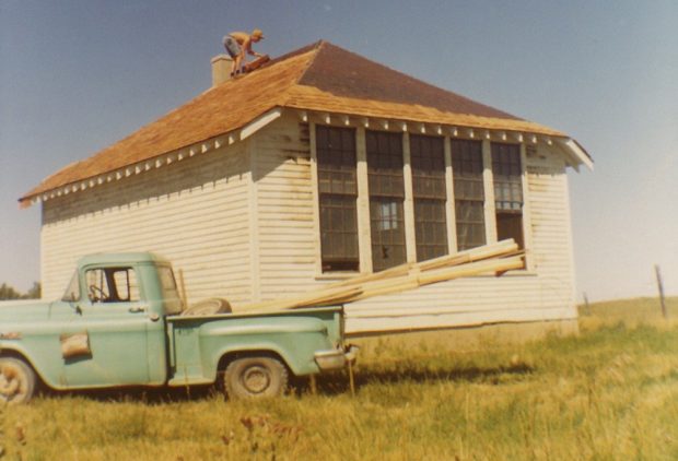 Photo couleur d’un homme travaillant sur la toiture d’une ancienne école blanche, avec une camionnette verte stationnée devant