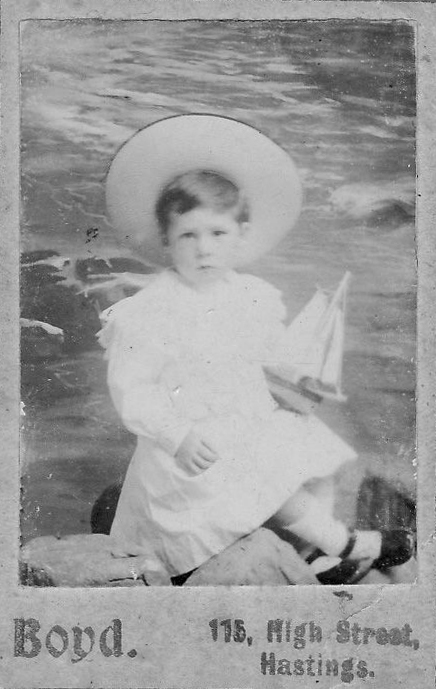 Portrait de studio en noir et blanc d’un jeune garçon tenant un bateau jouet et portant un grand chapeau et une robe blanche.