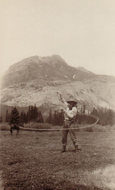 Photo sépia d’un homme en chapeau avec un lasso devant une montagne et un cheval au loin.