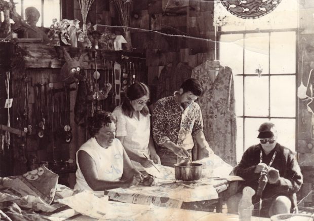 Photo noir et blanc de trois femmes et d’un homme travaillant dans un atelier d’art rempli de textiles et de céramiques; fenêtre lumineuse derrière.