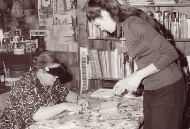 Photo noir et blanc d’une femme travaillant à une table avec une jeune étudiante en face; étagère et fournitures artistiques derrière.