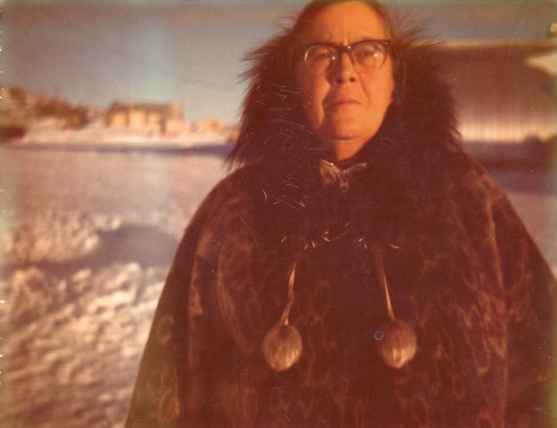 Photo couleur d’une femme avec des lunettes en manteau de fourrure, avec la ville enneigée en arrière-plan.