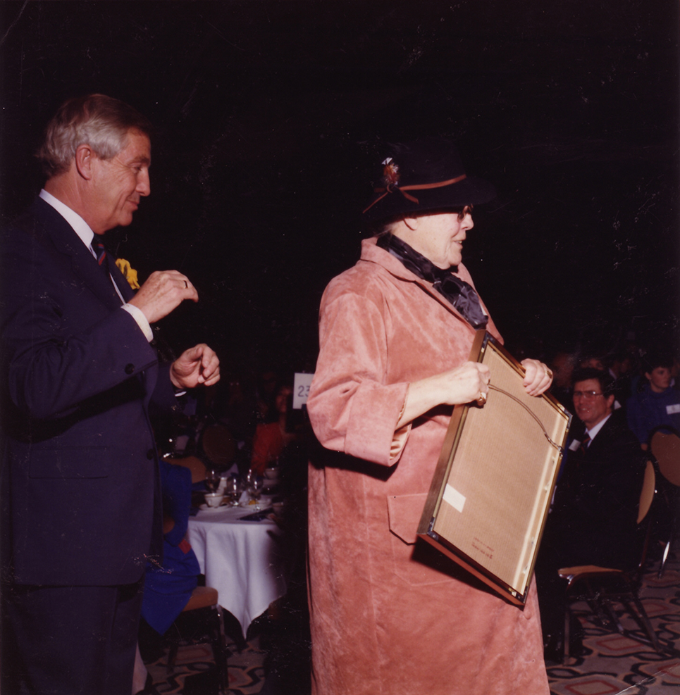 Photo couleur d’une femme portant un chapeau et une veste en daim, tenant un cadre; un homme en costume se tient derrière elle et une foule de personnes assises à des tables regardent.