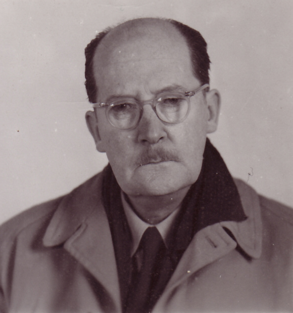 Photo noir et blanc d’un homme âgé avec des lunettes et une moustache, portant manteau et cravate.