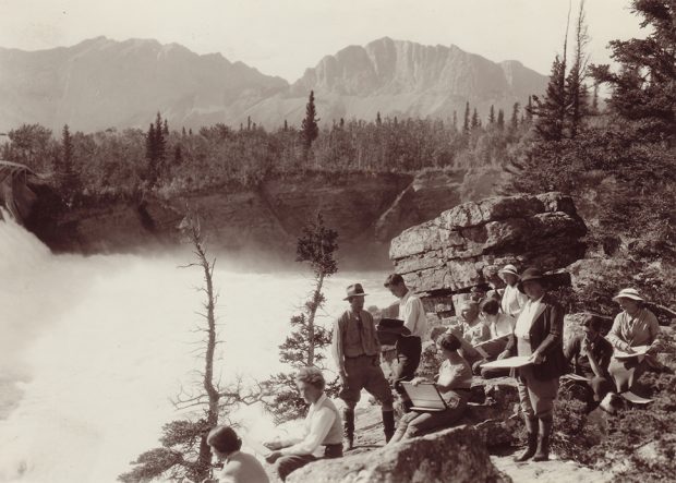 Photo sépia d’un groupe d’artistes faisant des croquis sur la rive surplombant une chute d’eau dans un paysage d’arbres et de montagnes.