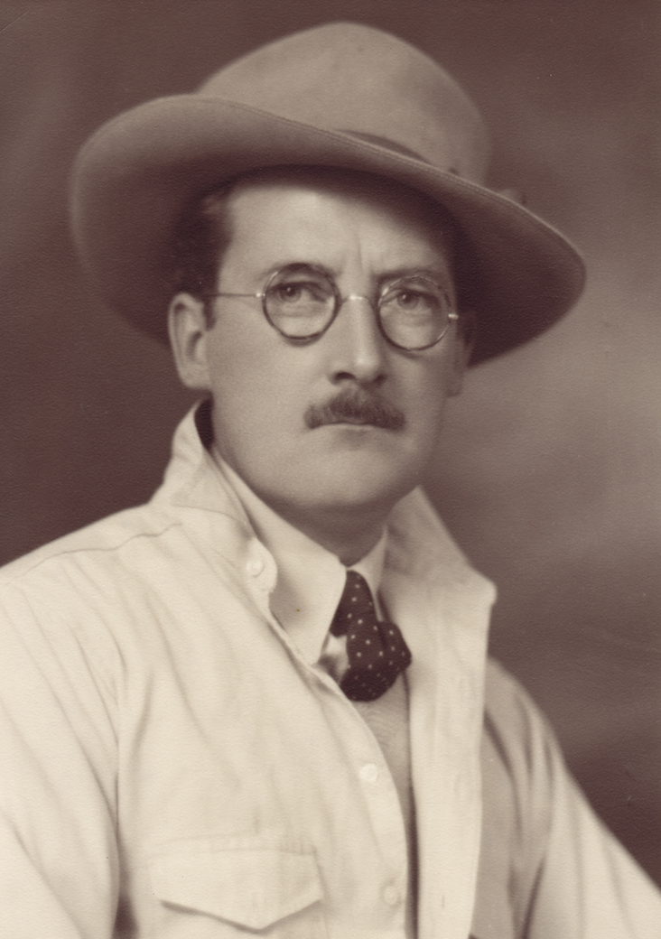 Portrait de studio sépia d’un homme moustachu portant des lunettes, un feutre, une cravate et une veste de couleur claire.