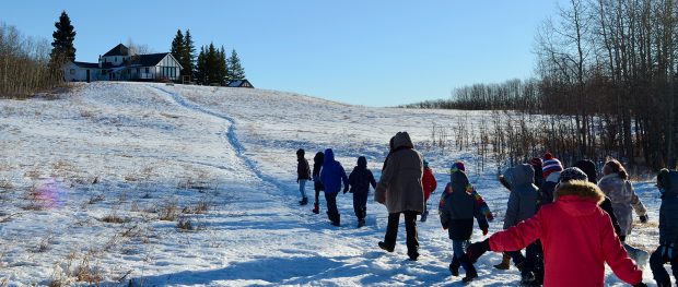 Photo couleur d’une file d’enfants marchant l’un derrière l’autre sur une colline dans un paysage hivernal vers une maison à colombages.