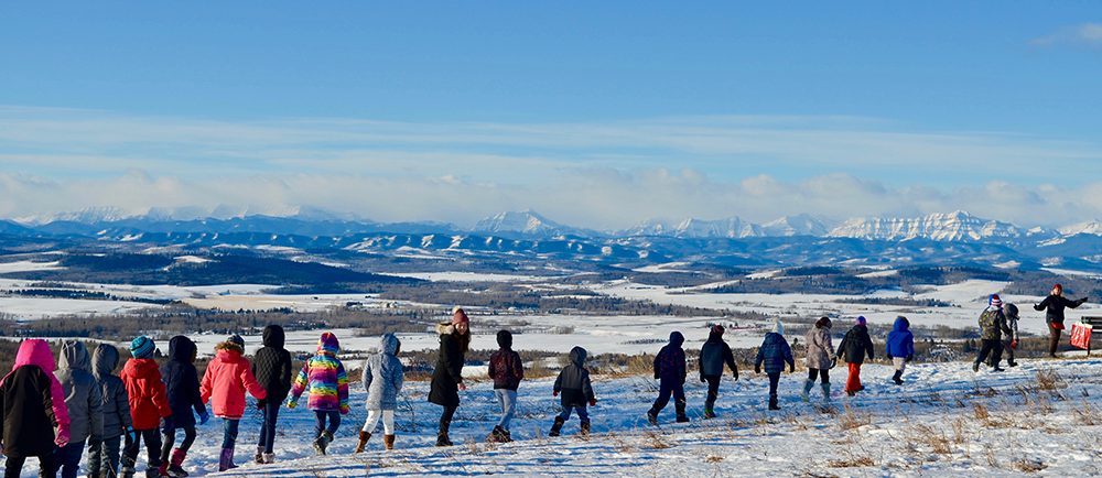 Photo couleur d’une file d’enfants marchant l’un derrière l’autre devant un panorama hivernal de montagnes dans le lointain.