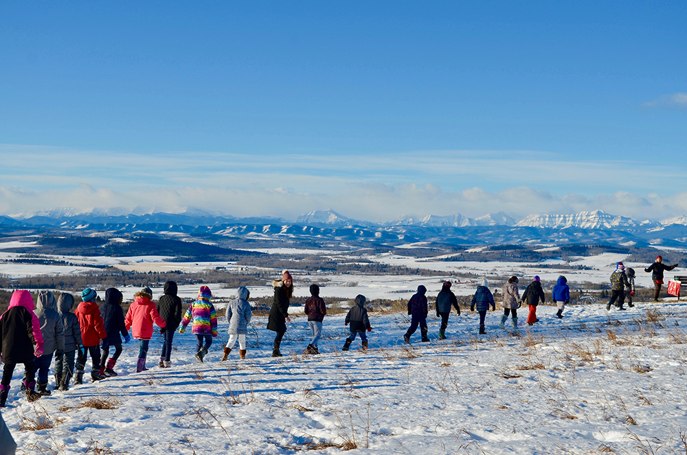 Photo couleur d’une file d’enfants marchant l’un derrière l’autre devant un panorama hivernal de montagnes dans le lointain.