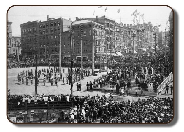 Image en noir et blanc de la très populaire célébration au centre-ville de Saint John, au Nouveau-Brunswick, en 1867, au retour de l’équipage de Paris, après la victoire à Paris. L’intersection des rues King et Water est bordée de milliers de gens. Dans la zone ouverte, au centre de la place, des activités de célébration sont offertes aux spectateurs. Des estrades ont également été aménagées en face de la place centrale. Des décorations ornent les façades des magasins et des guirlandes de mini-drapeaux triangulaires sont suspendues entre les poteaux téléphoniques et les lampadaires.