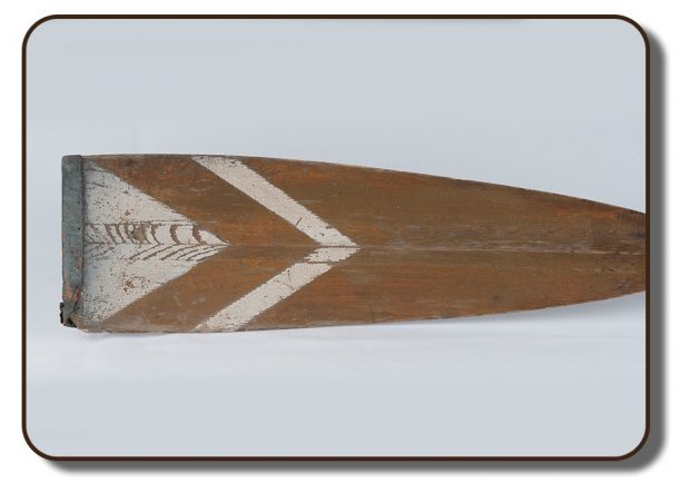 Image d’une rame en bois qui aurait été utilisée par les membres de l’équipage de Paris. À l’extrémité de la palette, il y a un ornement décoratif de forme triangulaire peint en blanc. Il y a aussi un revêtement de cuir sur le bout de la palette, qui est endommagé et s'effrite à l’une des extrémités en raison de l’usage répété ou de l’âge.