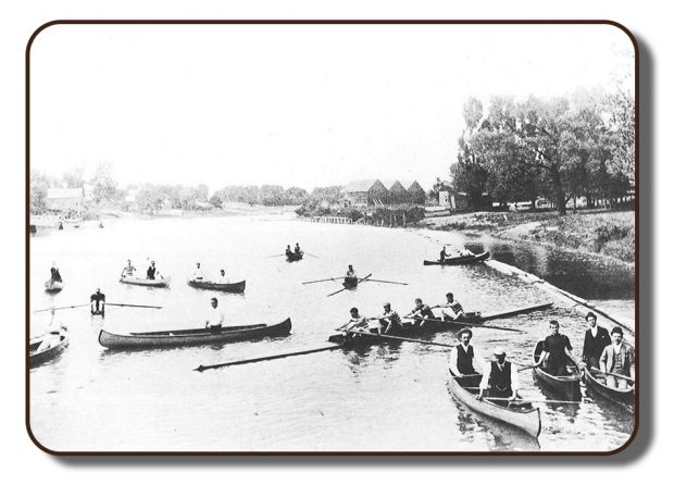 Image d’une berge dans la région de Renforth, à Rothesay, Nouveau-Brunswick, avant la participation de l'équipage de Paris à la Régate internationale d’aviron, en juillet 1867. Cette photographie est en noir et blanc; elle a pâli au fil des années et en raison de l'équipement de photographie personnelle disponible à cette époque. Il y a de nombreuses embarcations sur le cours d’eau, y compris l’aviron à quatre rameurs de l’équipage de Paris, des rameurs seuls dans leur embarcation respective et des canoës comptant une ou deux personnes. Le long de la berge, il y a une zone boisée et on voit ce qui semble être trois granges ou hangars à bateaux.