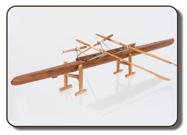 Image d’un modèle d’un des premiers avirons à deux rameurs. Ce modèle est fait en bois et mesure environ 10 pouces de long et comprend quatre rames en bois fixées par des portants métalliques.