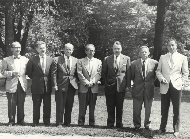 Photographie noir et blanc de sept hommes debout à côté de l’autre, à l’extérieur. 